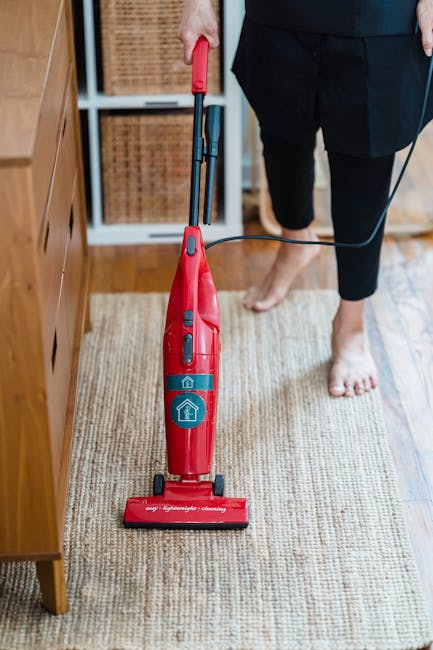 Two individuals are engaged in surface cleaning of a rolled-up beige carpet with blue straps, positioned on a wooden floor in a living room. The woman, with curly brown hair and wearing a green shirt,